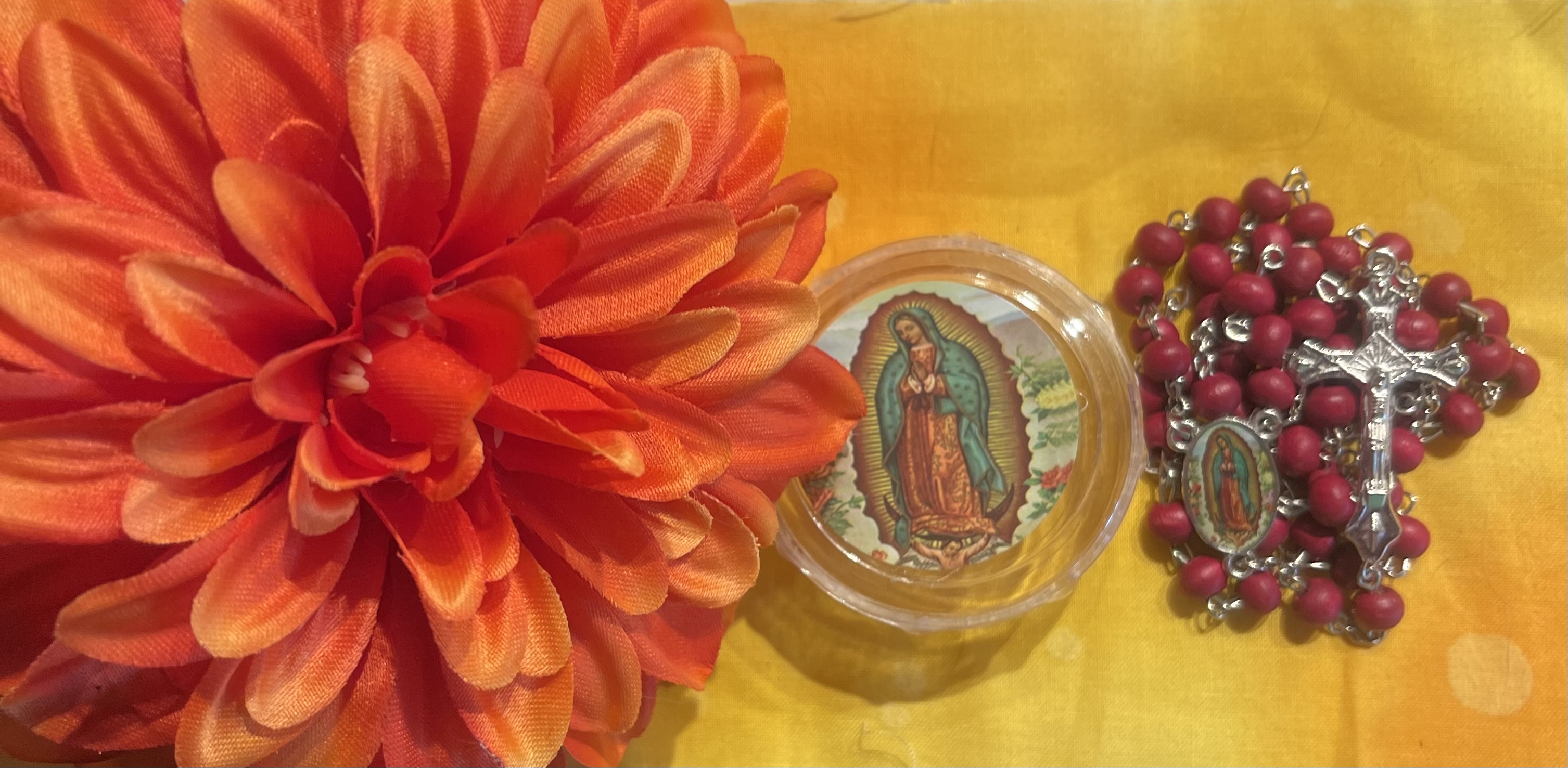 Bright orange flower by a round case showing the Virgin of Guadalupe next to a rosewood rosary